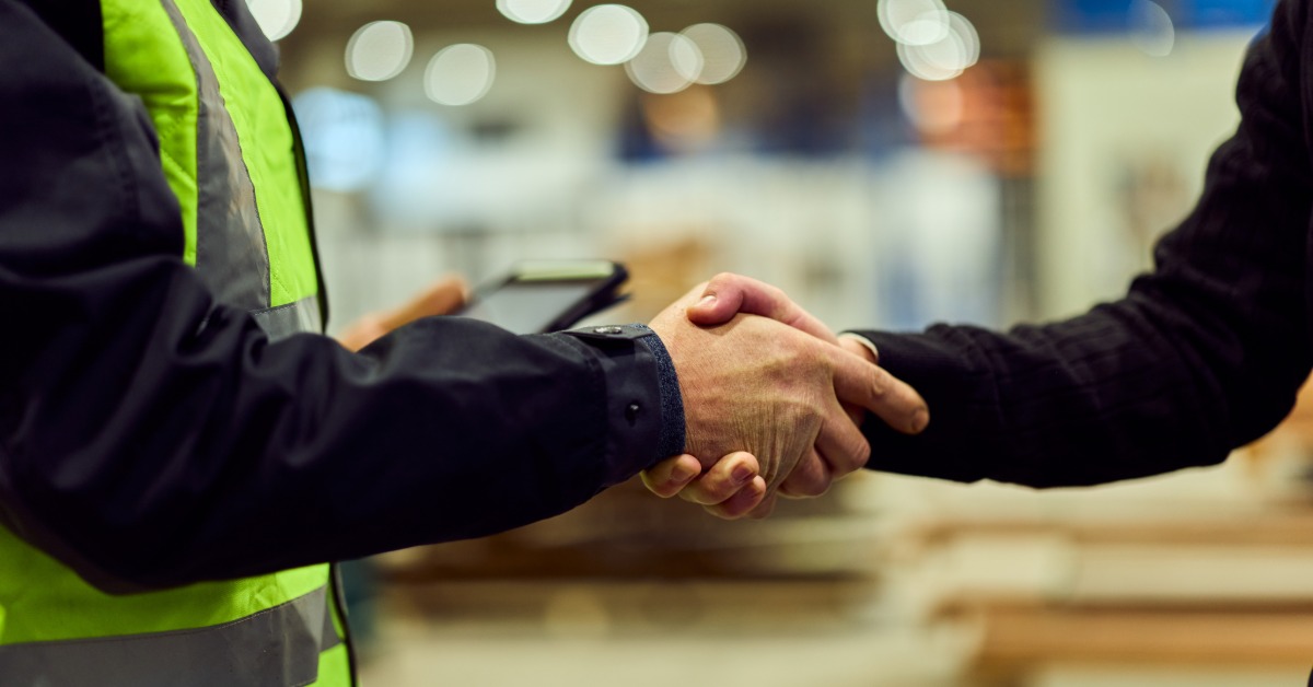 A manufacturing team employee in high-visibility gear shaking hands with another professional in a dress shirt.