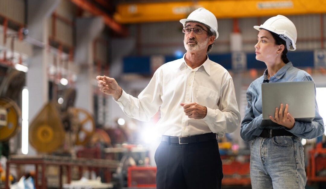 Two professionals walking side by side in a manufacturing plant floor while wearing white hard hats.