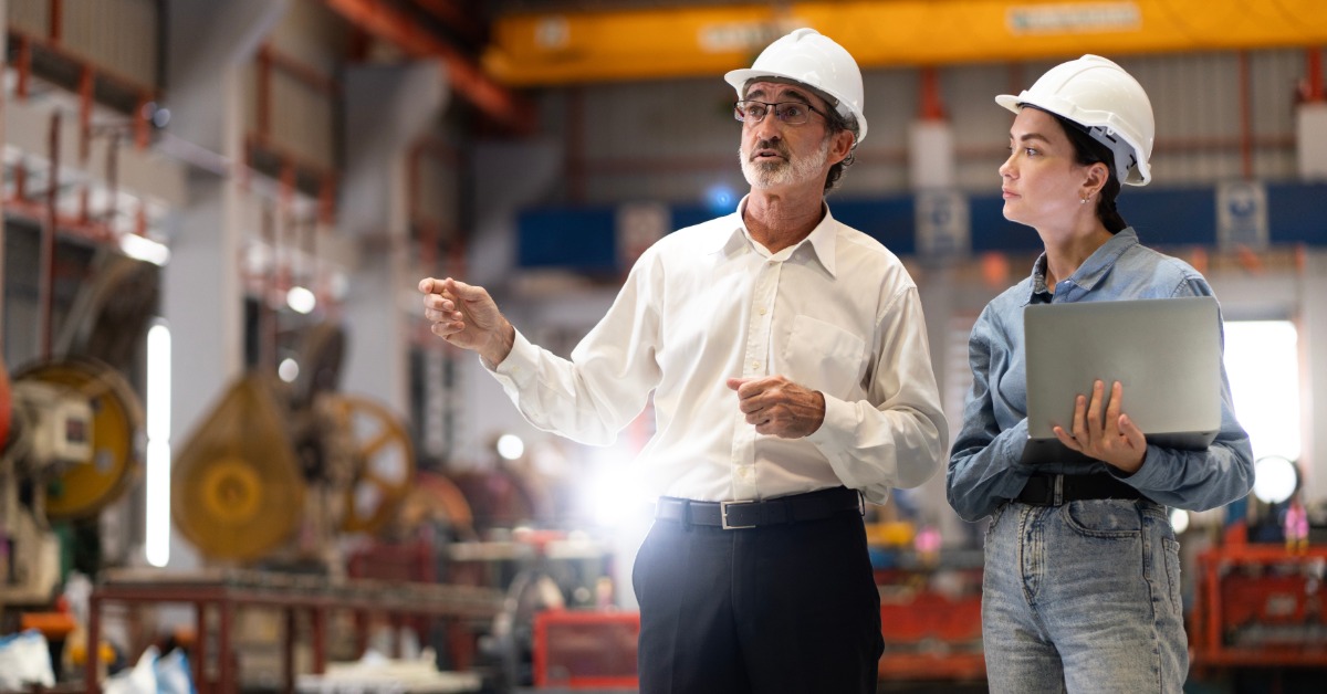 Two professionals walking side by side in a manufacturing plant floor while wearing white hard hats.